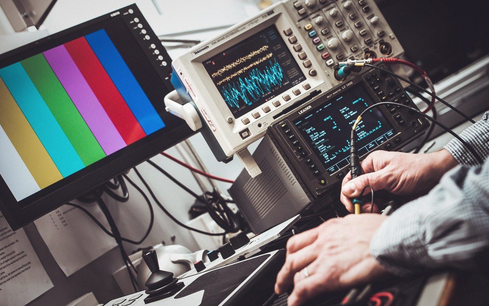Technician's hands adjust equipment beside an oscilloscope and color bar test monitor